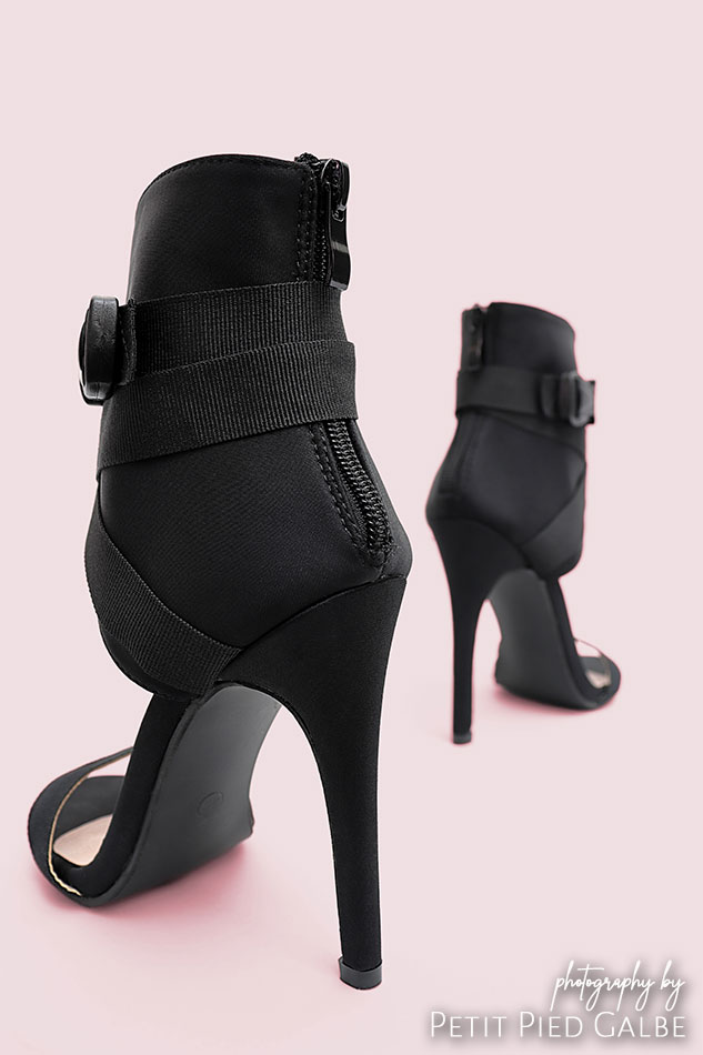 Black heeled sandals showcased from behind on solid pink background.