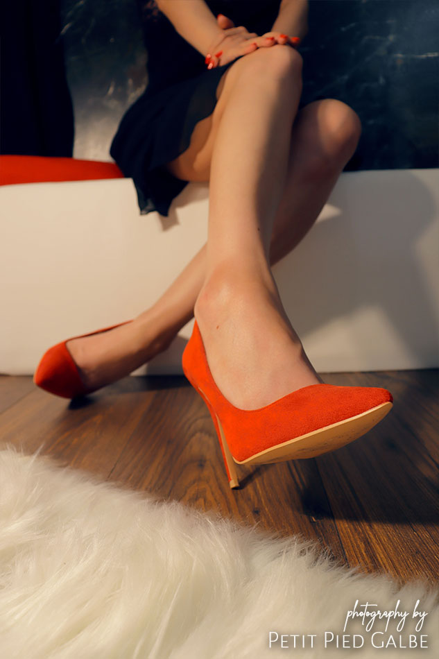 Elegant lady in red high heels and black dress sitting on a white pouf in stylish interior. Woman is showing shoes close to the camera.