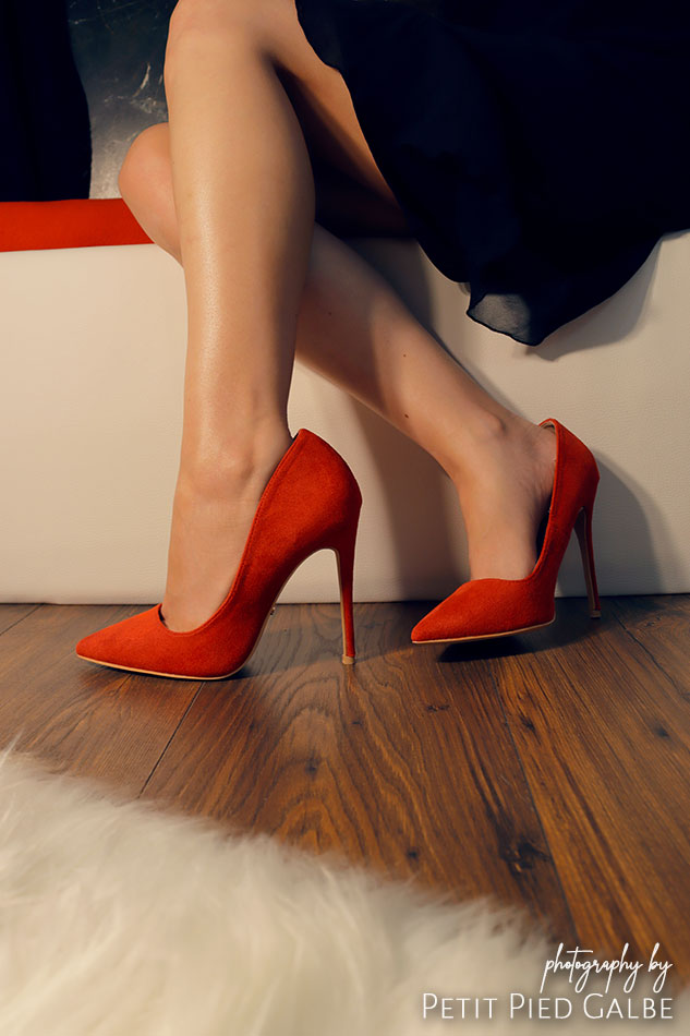 Beautiful lady in red high heels and black dress sitting in elegant interior on a white sofa. Close-up on the shoes.